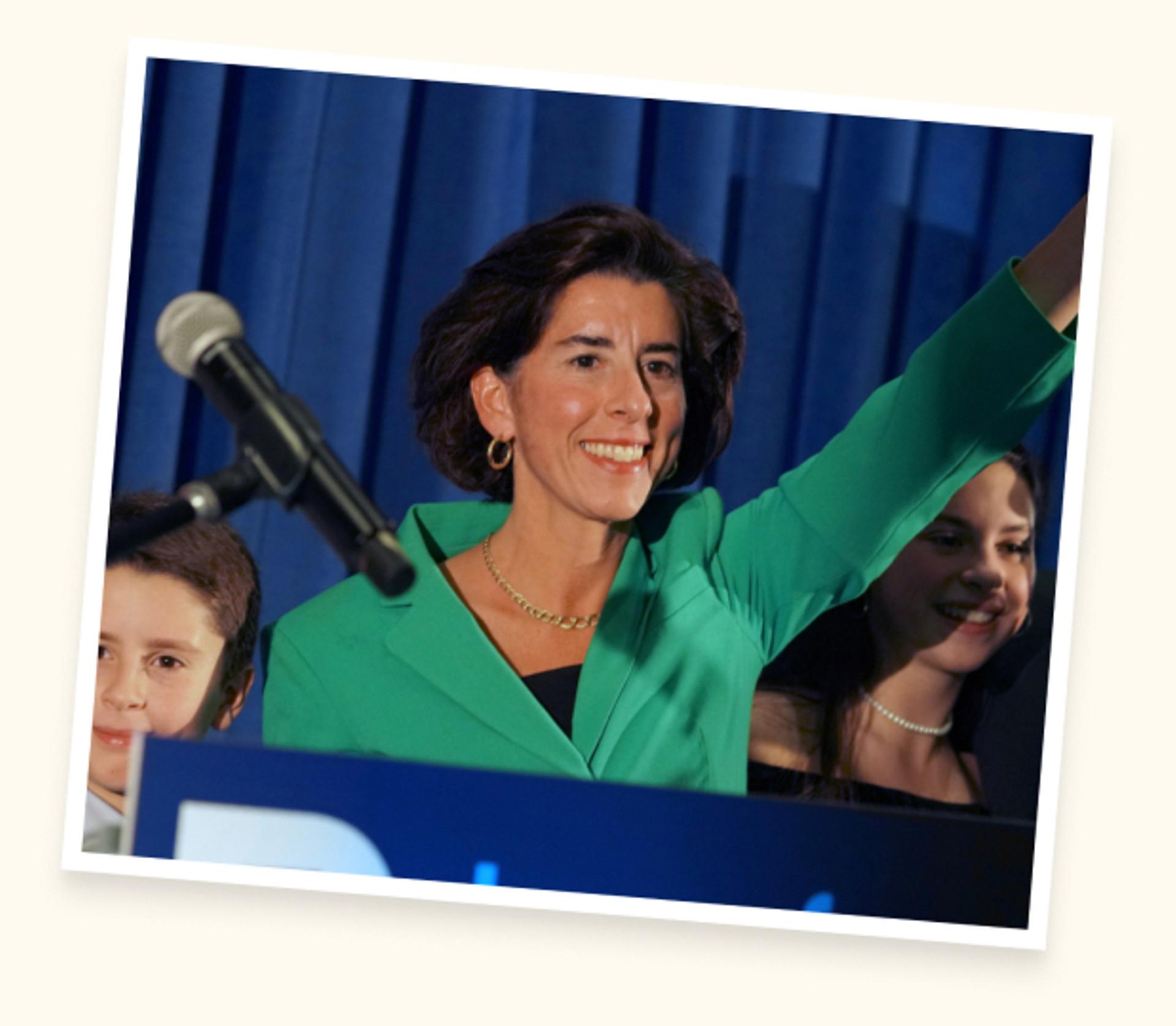 A photograph of Gina Raimondo standing a podium, smiling, and raising her arm.