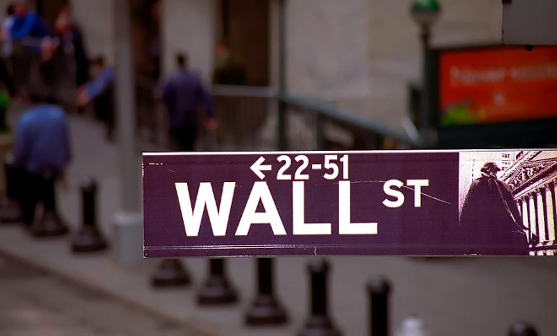 A photograph of a street sign of Wall St. People walk by and part of the Subway entrance is visible in the background.